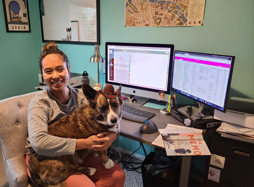 Karina with a corgi wearing a hat sitting on her lap in front of a desk with two monitors.